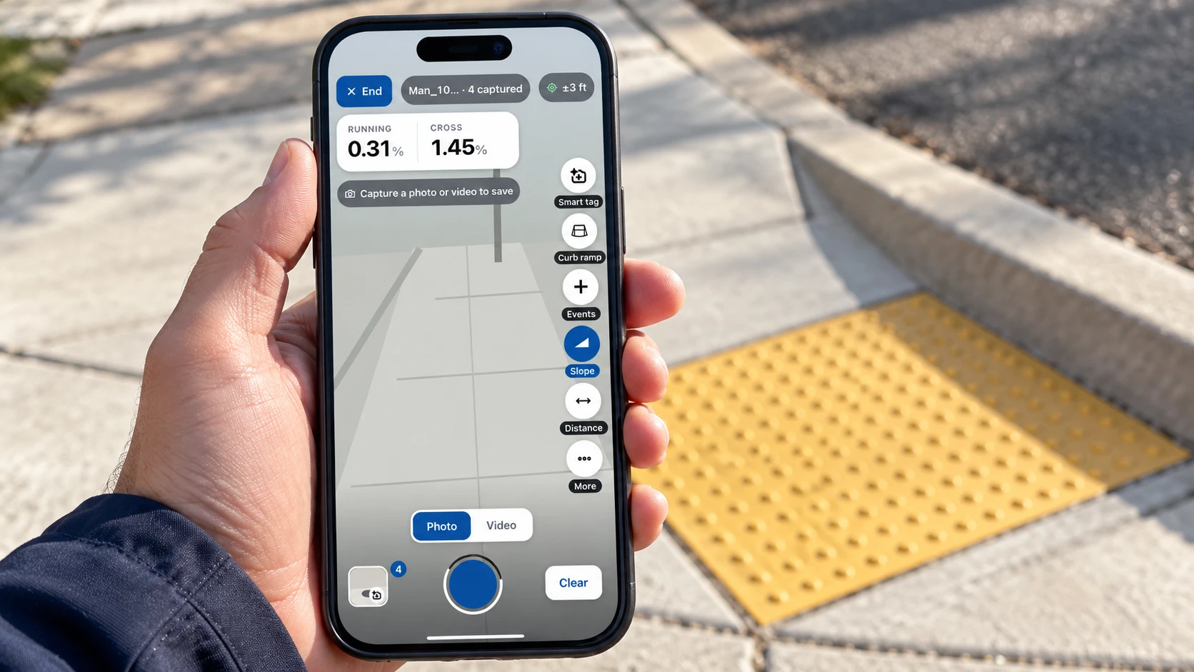 A hand holding a phone at a curb ramp, with the ADAM camera view showing running and cross slope readings